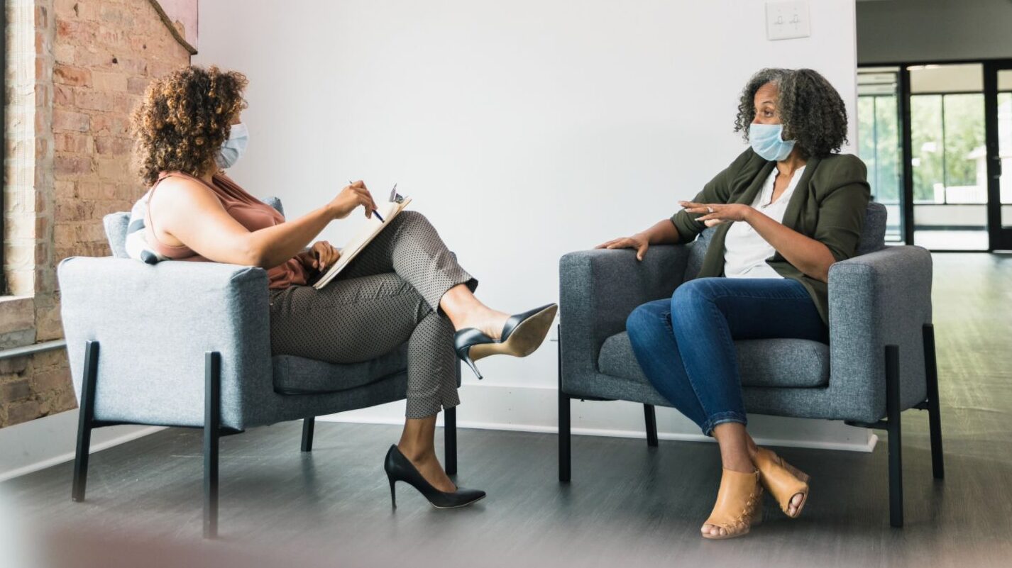 Two women with curly hair wearing face masks sit in perpendicular chairs. They are talking, and one has a clipboard.